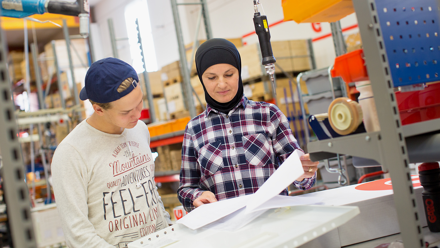 Kvinna i slöja visar dokument för ung man i keps i fabriksmiljö