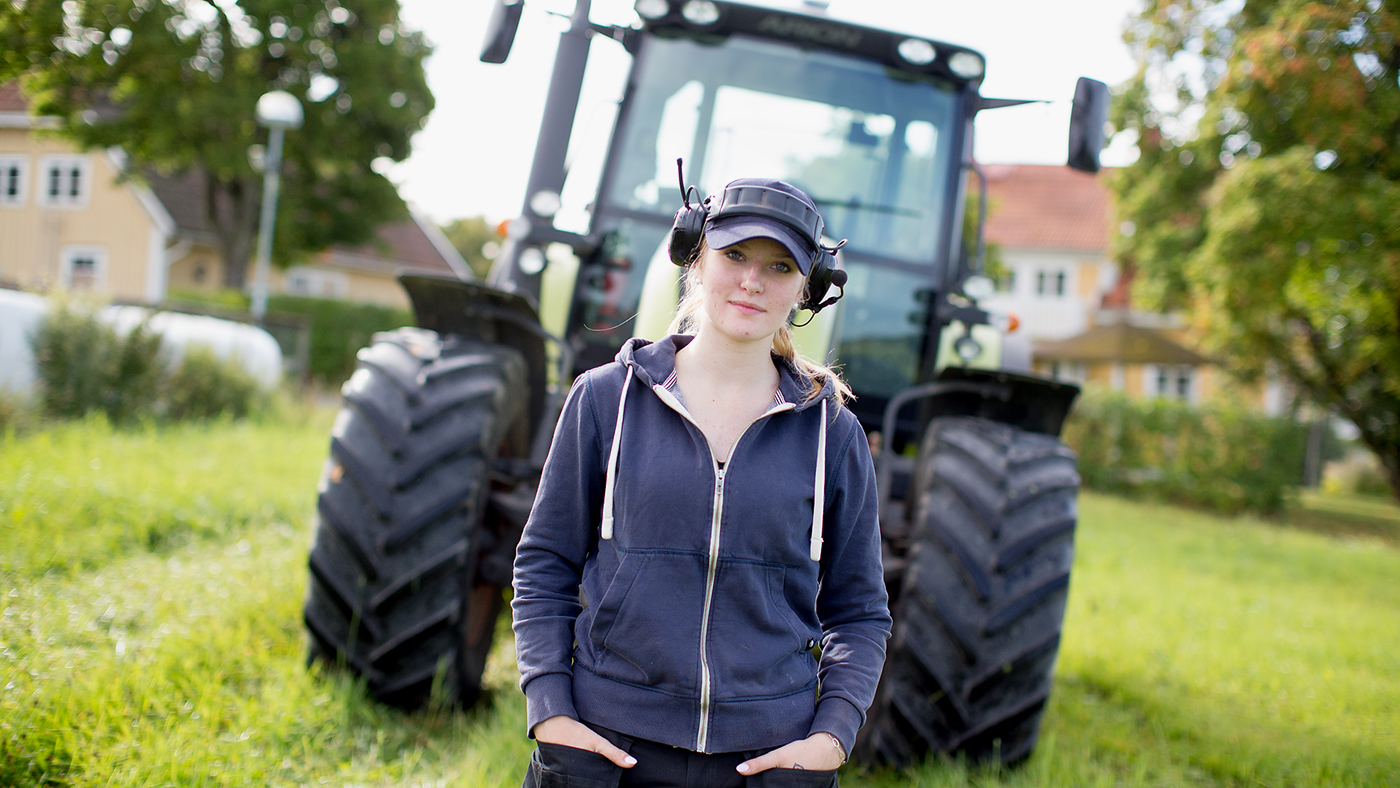 Ung kvinna står på åker framför en traktor. 